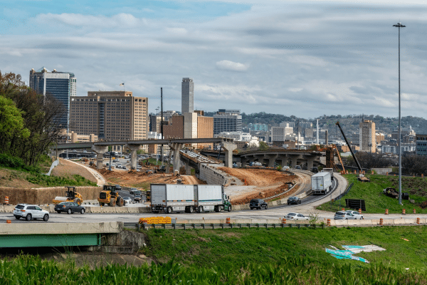 Highway interchange construction with cranes, traffic, and city buildings in the background, showing roadway infrastructure improvements in progress.