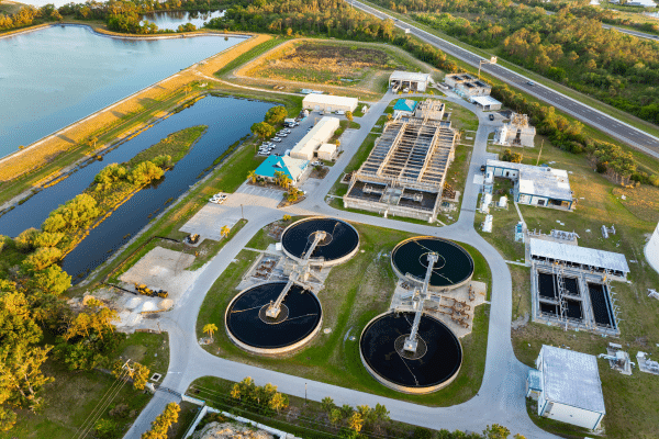 Aerial view of a municipal water and wastewater treatment facility with circular tanks, processing buildings, access roads, and nearby retention ponds.