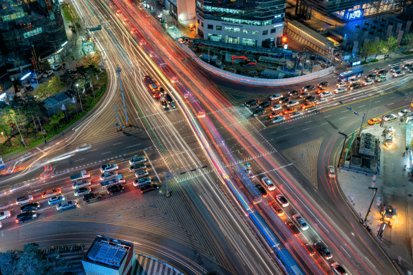 Aerial nighttime view of a busy city intersection with traffic light trails, roadway markings, vehicles, and urban transportation infrastructure.