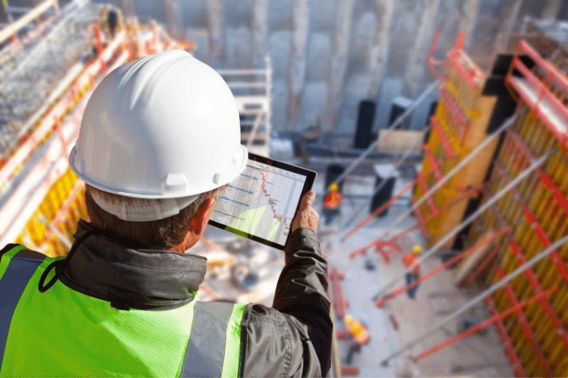 Construction professional using a tablet to review project plans at an active construction site with excavation work and structural supports visible in the background.